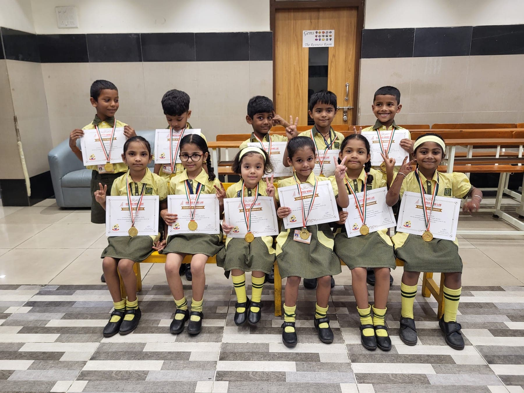 Eleven Kindergarten children in yellow and green uniform holding certificates of achievement, medals round their necks.