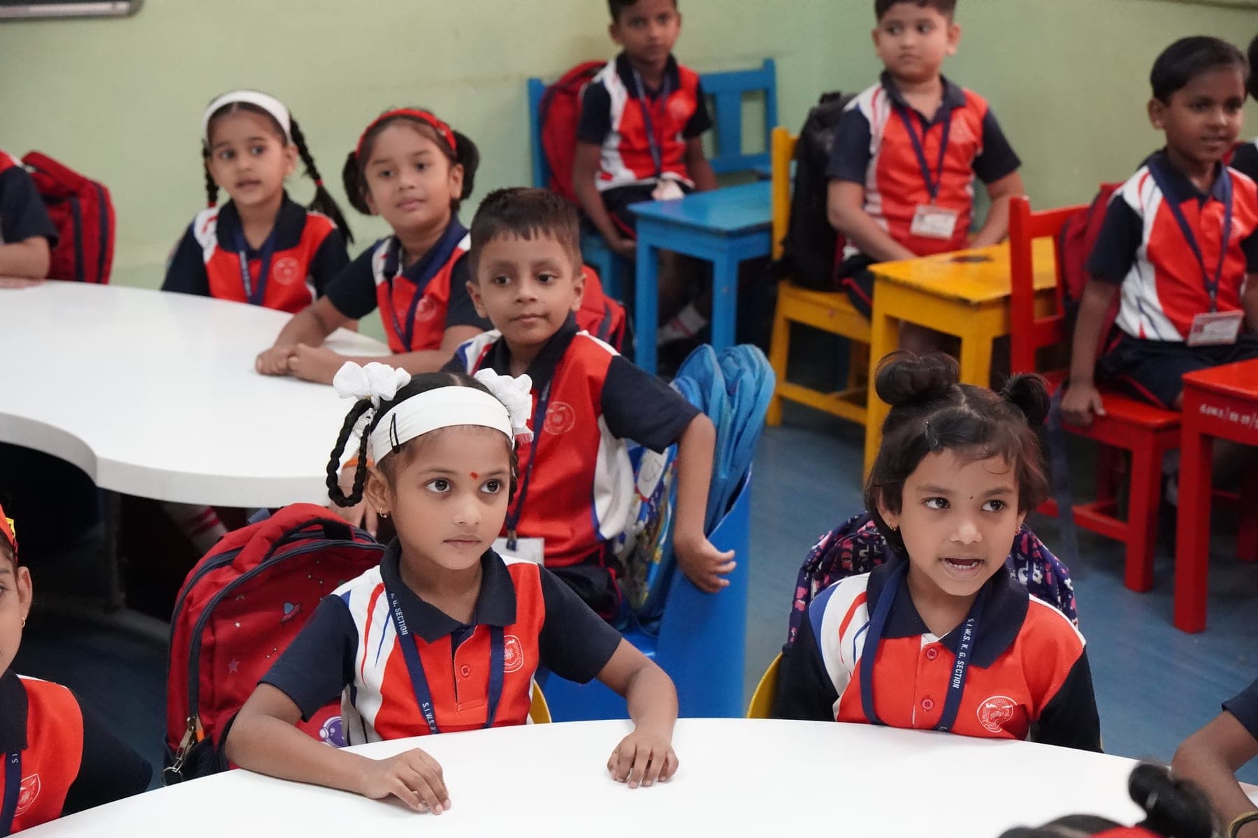 Young children in SIWS uniform seated at classroom tables, listening to their teacher.