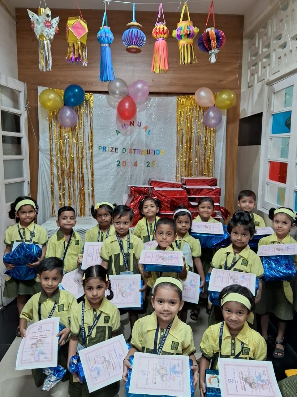 Kindergarten children in yellow and green uniform holding Certificates of Honour and wrapped prizes, under paper lanterns and a banner reading “Annual Prize Distribution 2024-25”.