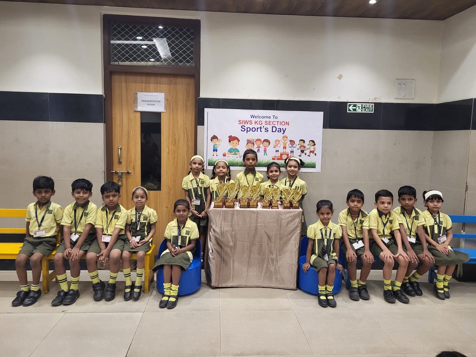 Kindergarten children in yellow and green uniform seated on benches along both sides of a draped table of trophies, beneath the sports day banner outside the presentation room.