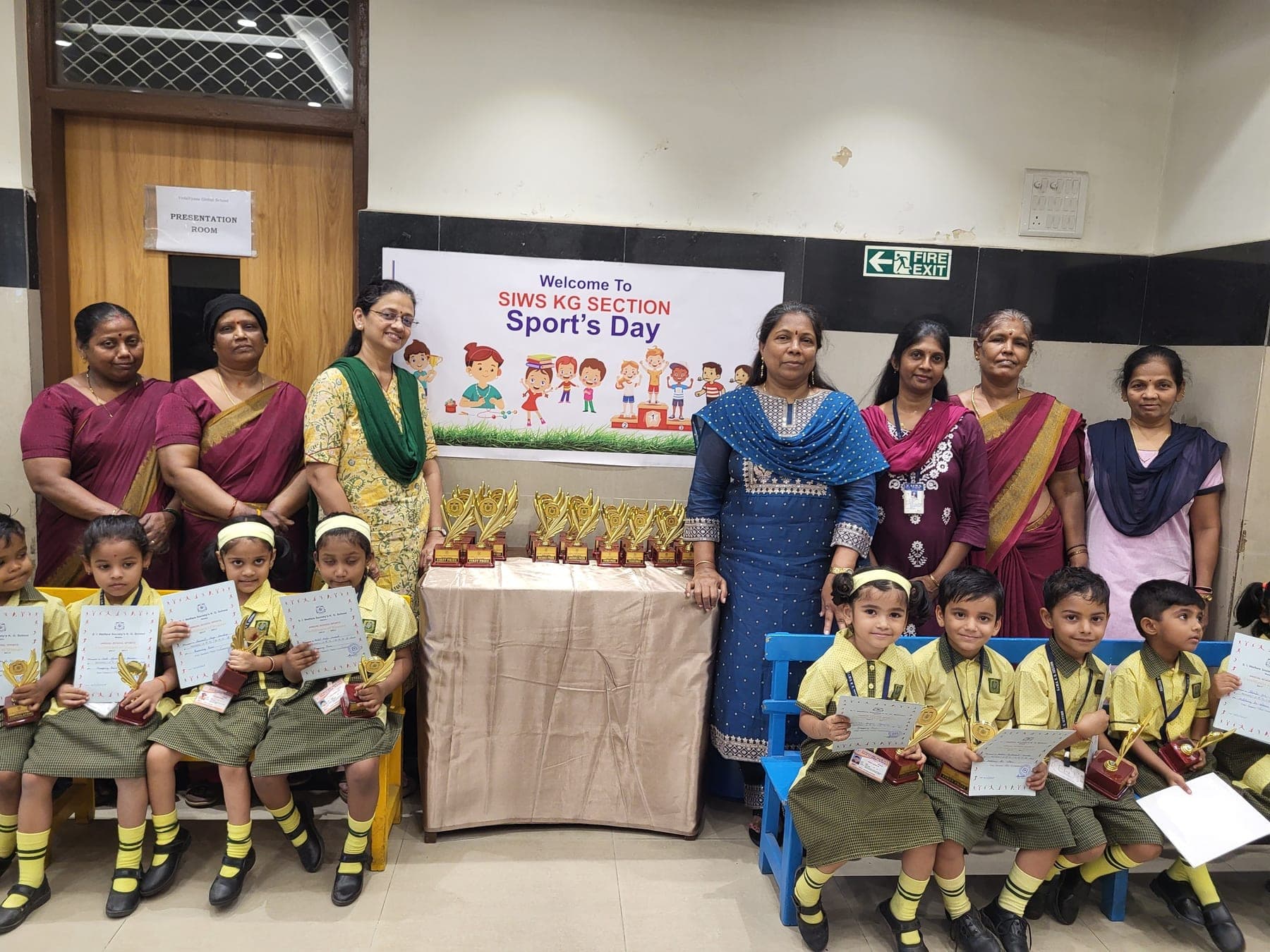 Seven Kindergarten staff standing behind a draped table of trophies beneath the sports day banner, with children in yellow and green uniform seated on benches to either side holding certificates.
