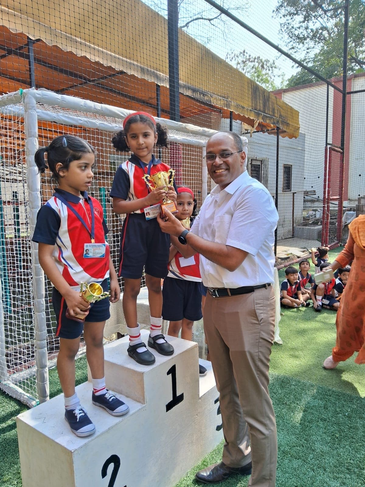 A man in a white shirt and tie handing a gold trophy to a girl standing on the number 1 block of the winners’ podium, with two more Kindergarten children on the second and third blocks, on the green turf pitch.