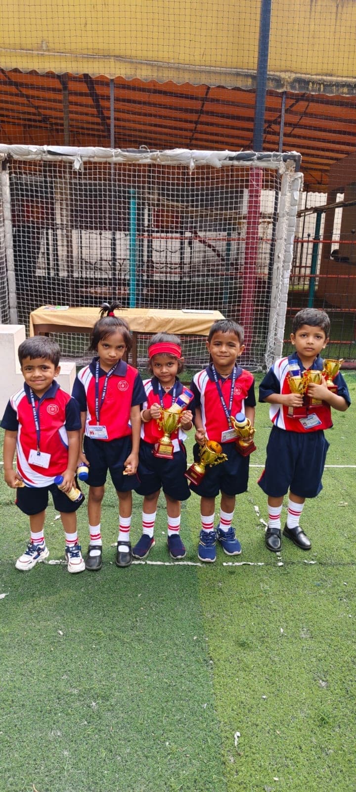 Five Kindergarten children in red and navy sports kit standing in a line on the turf pitch, each holding a gold trophy.