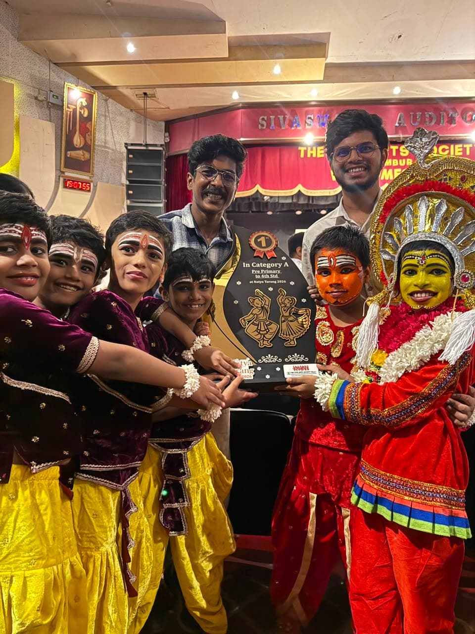 Children in dance costume and face paint holding a trophy reading “1st in Category A, Pre-Primary to 4th Std at Natya Tarang 2026”, with two teachers behind them.