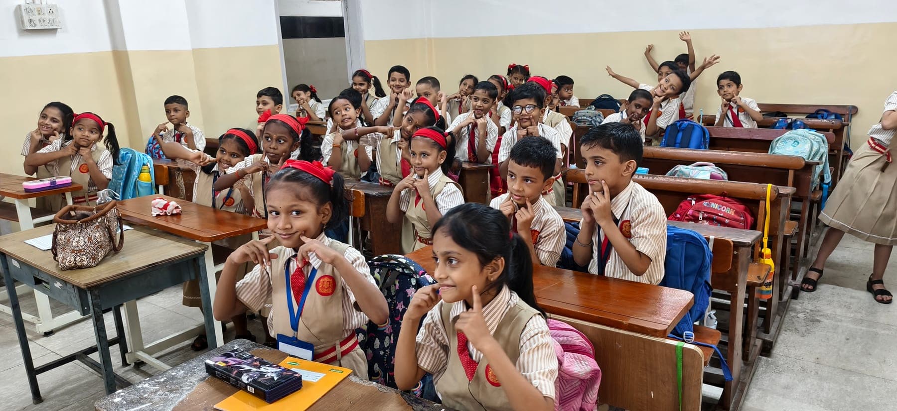 A full Primary class in striped uniform at their bench desks, turning towards the camera with their teacher’s room around them.