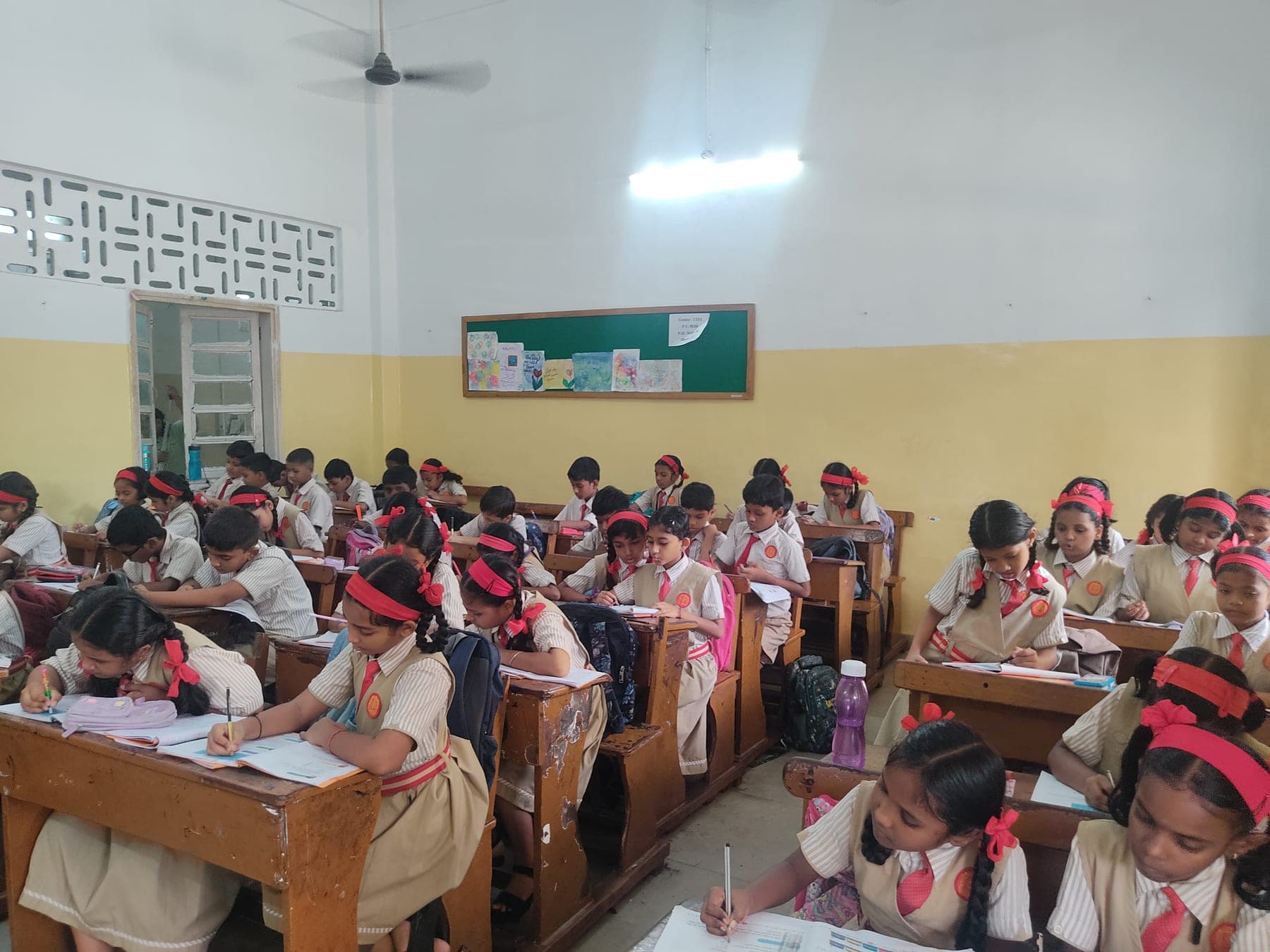 A full Primary classroom of children in cream uniform with red ties and hairbands, working at wooden double desks under a high yellow-and-white wall.
