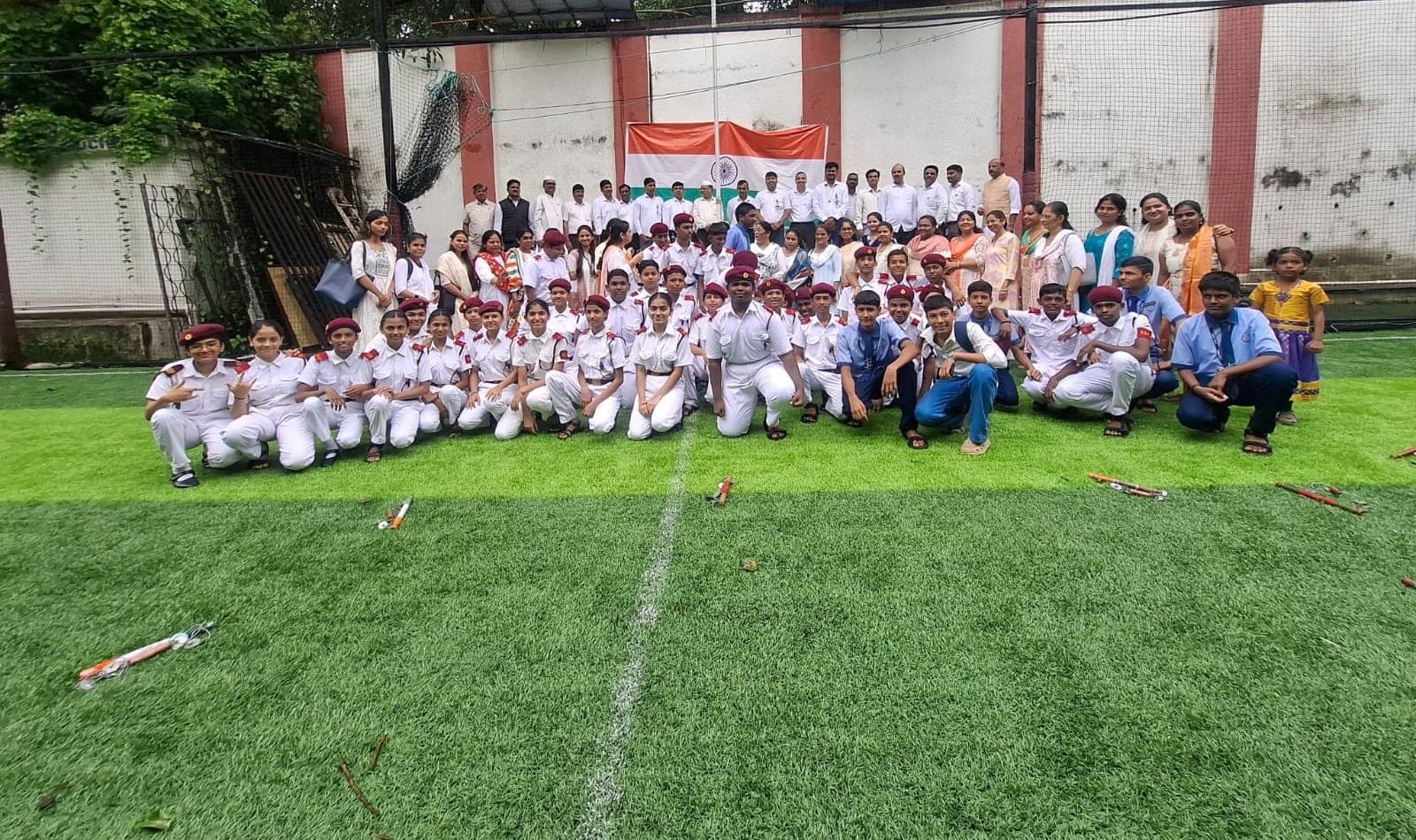 Students in ceremonial white uniform and maroon berets kneeling and standing in rows on the college ground beneath an unfurled national flag, with staff and families behind them.