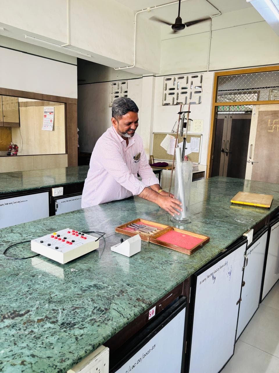 A member of the physics department at a laboratory bench setting up a measuring cylinder and apparatus, with a resistance box and a case of instruments laid out beside him.