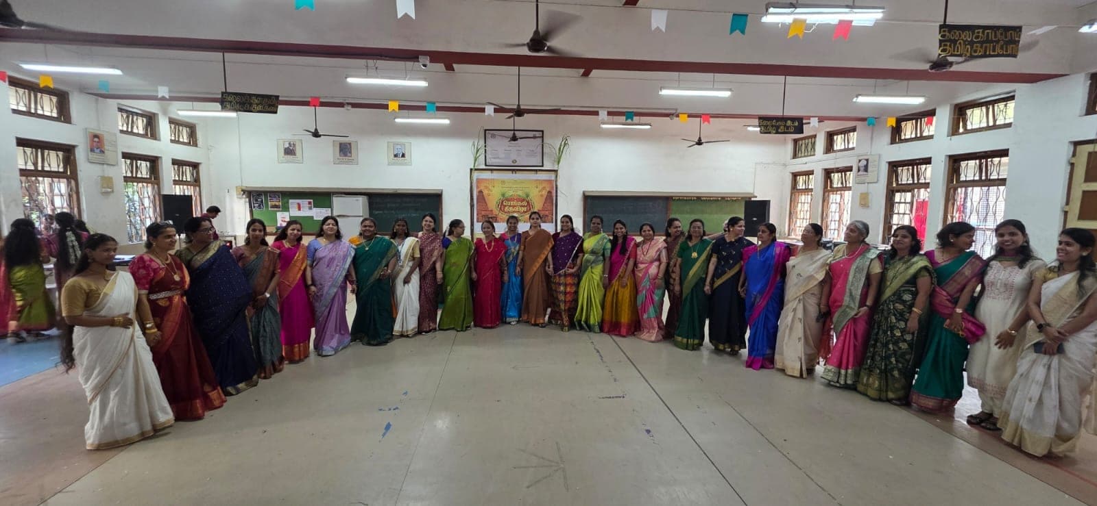 Staff of the college standing in a long line across the hall in bright silk sarees, beneath Tamil banners and bunting, with sugarcane framing a Pongal Thiruvizha backdrop.