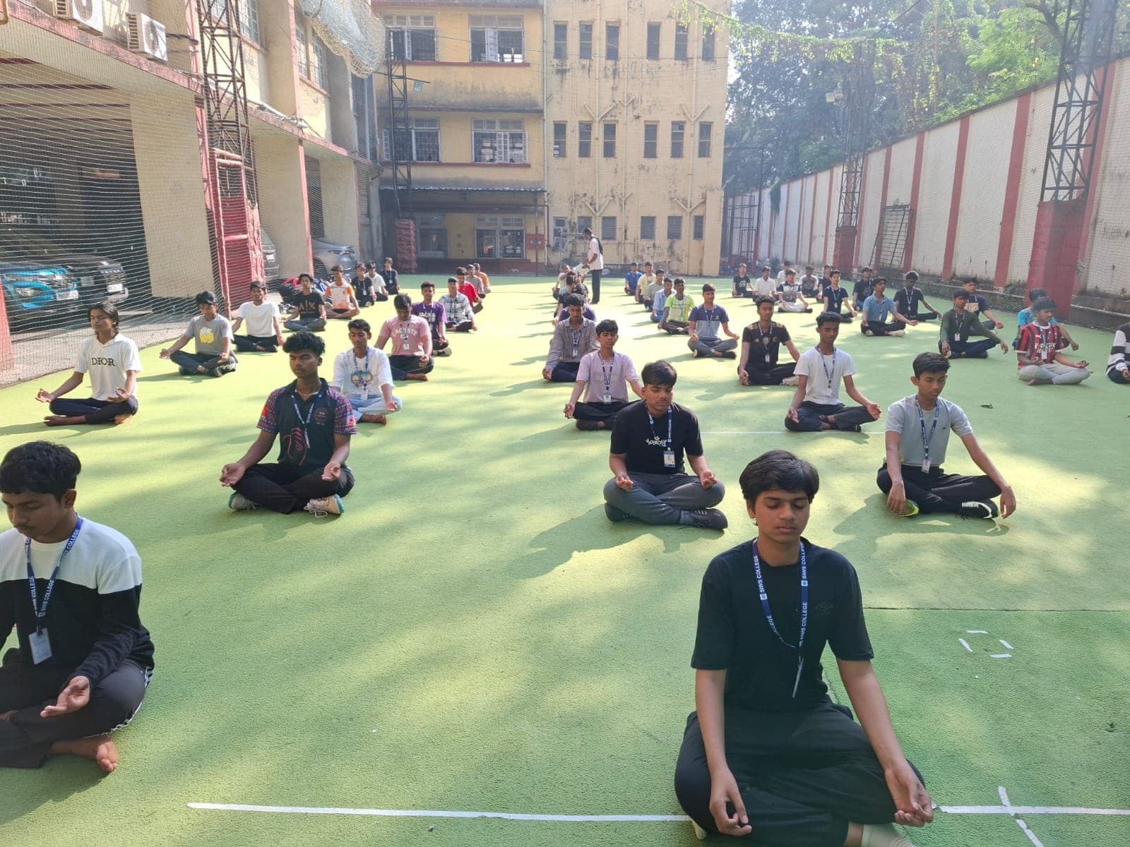 Junior College students seated cross-legged in rows on the open ground with their eyes closed and hands resting on their knees, during a guided meditation.