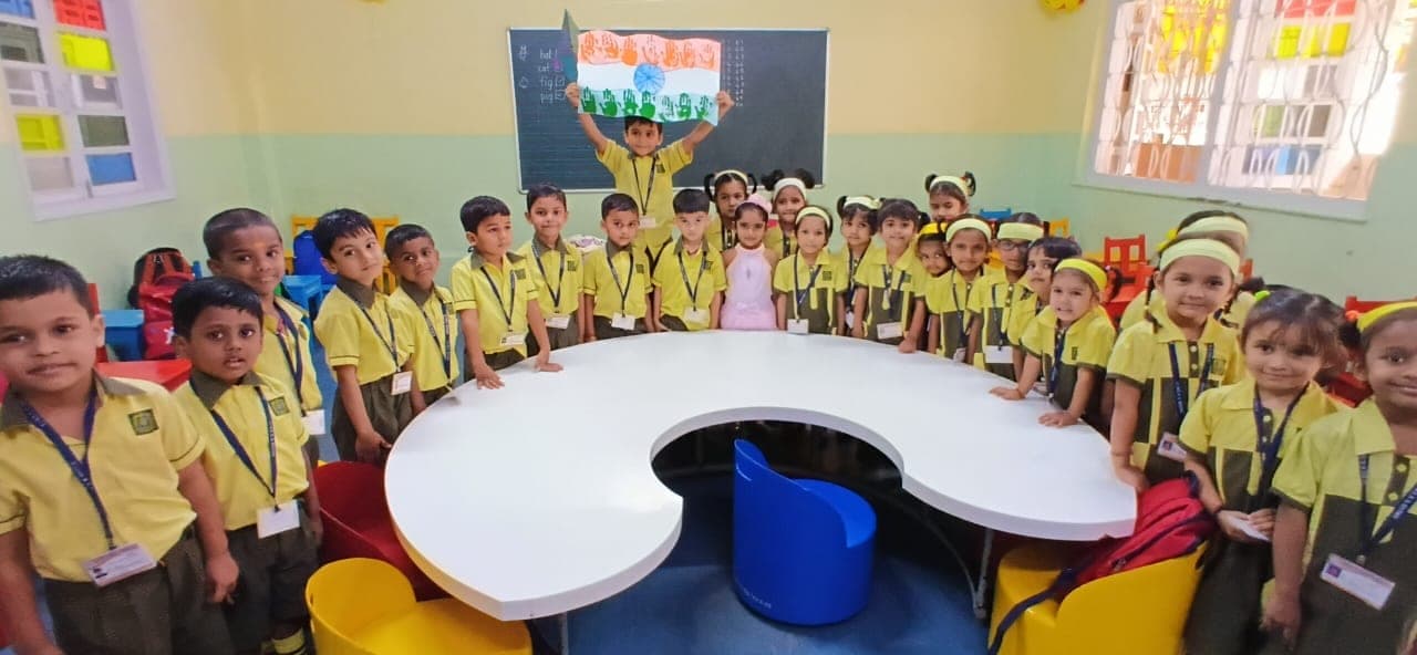 A full kindergarten class in yellow and green SIWS uniform standing all the way around a large curved white table, with a child at the back holding up a handmade Indian flag made from painted handprints.
