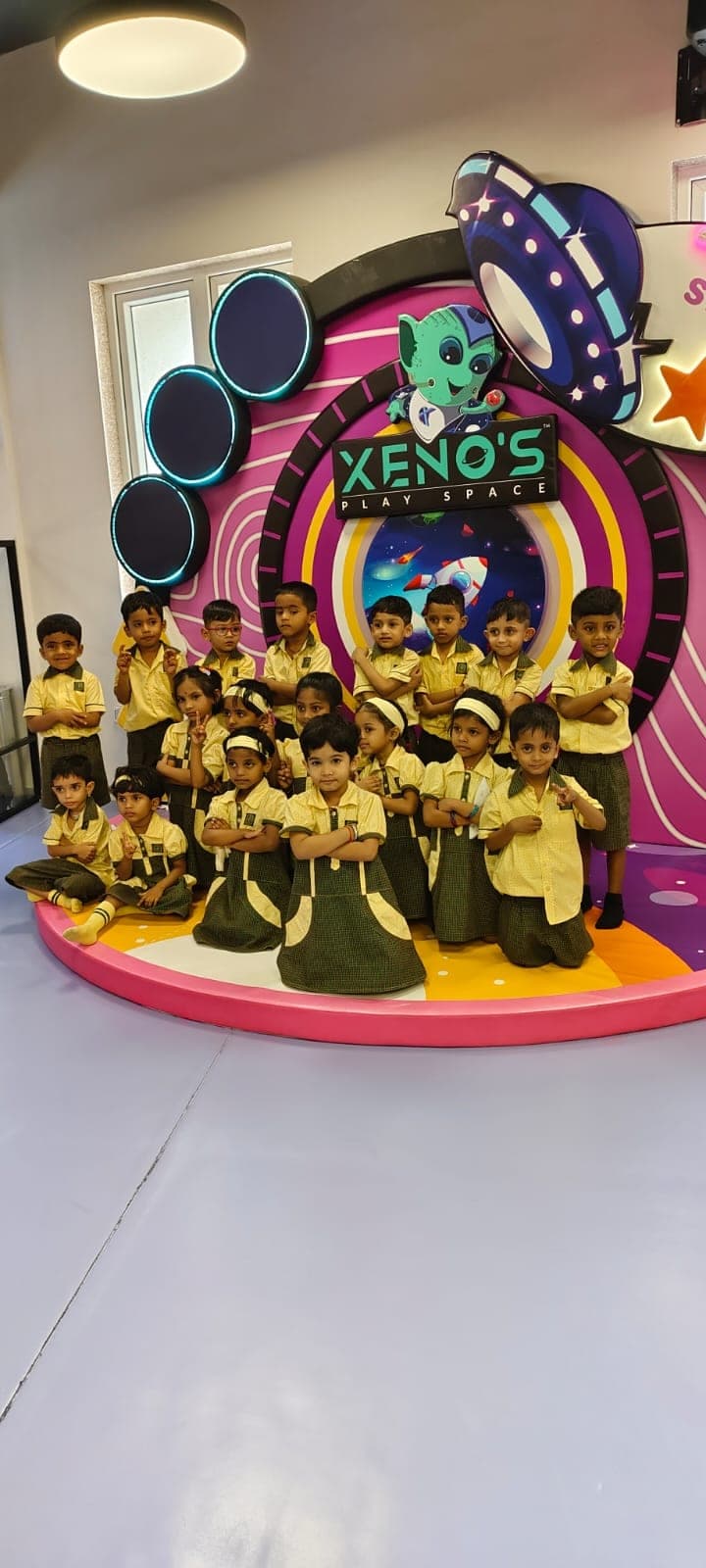 A kindergarten class posing together on a low round stage in front of a brightly lit space-themed backdrop at an indoor play centre, during a class outing.
