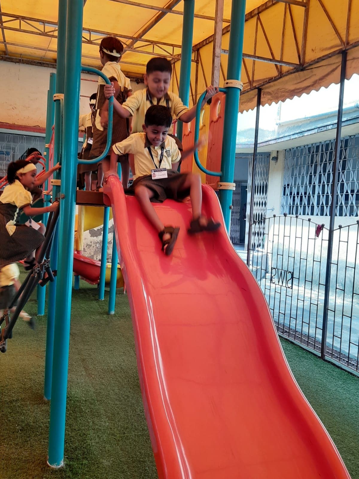Kindergarten children playing on a red slide and climbing frame under a canopy, on the green artificial-turf play area, one child mid-slide and others waiting their turn on the platform above.