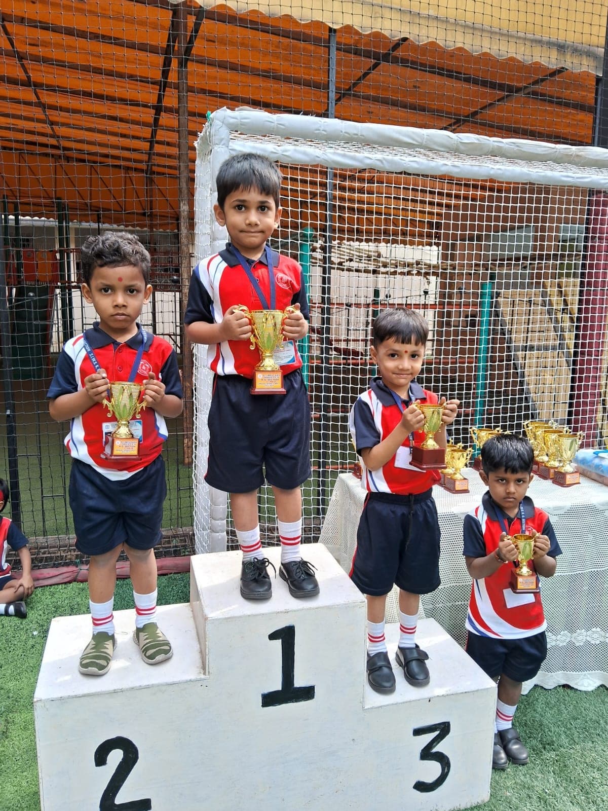 Four kindergarten children in red and navy sports kit on a winners’ podium numbered 1, 2 and 3, each holding a gold trophy, with a table of further trophies behind them on the turf pitch.