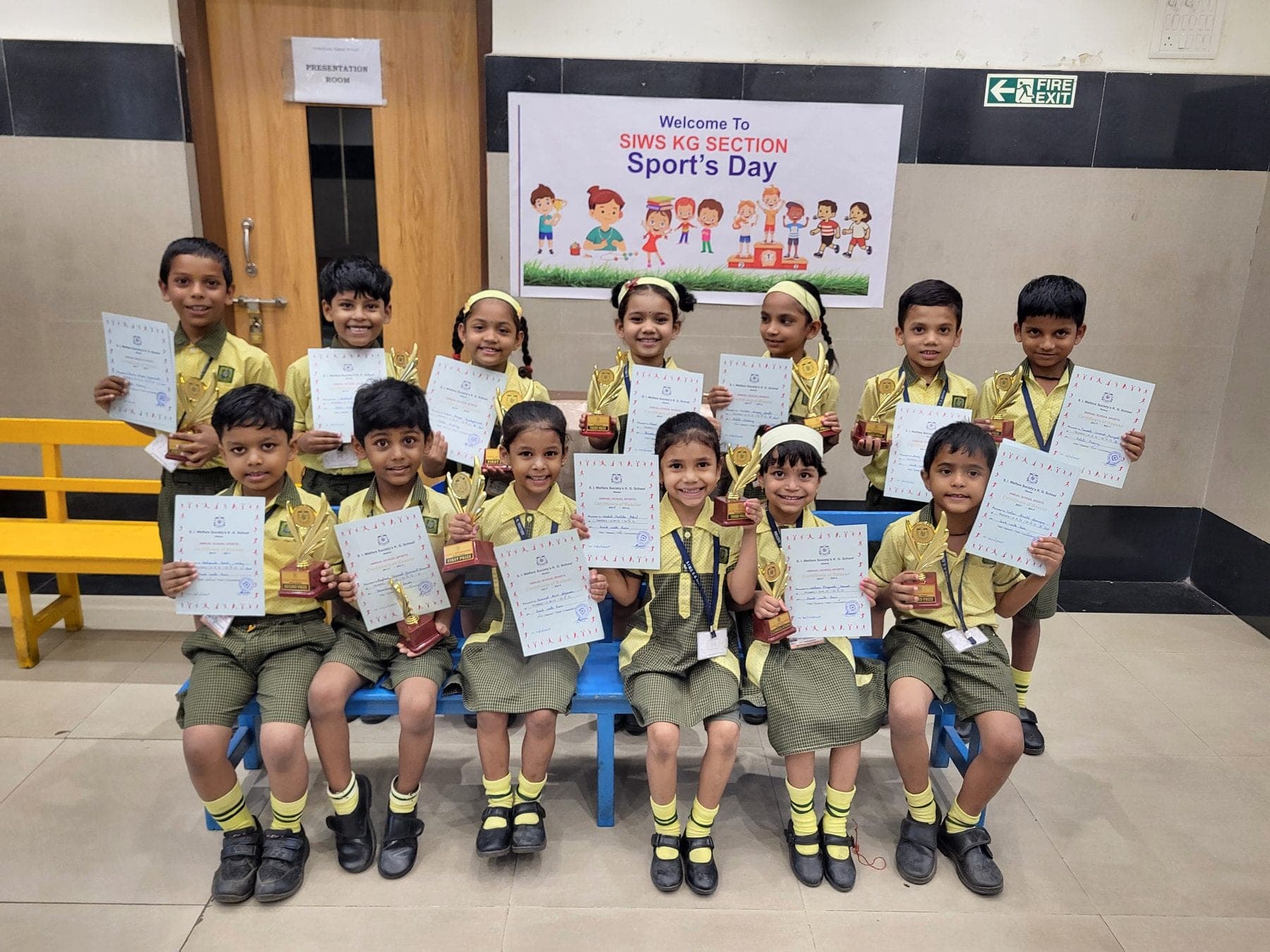 Thirteen kindergarten children holding certificates and trophies, seated and standing in two rows beneath a banner reading “Welcome to SIWS KG Section Sport’s Day”.