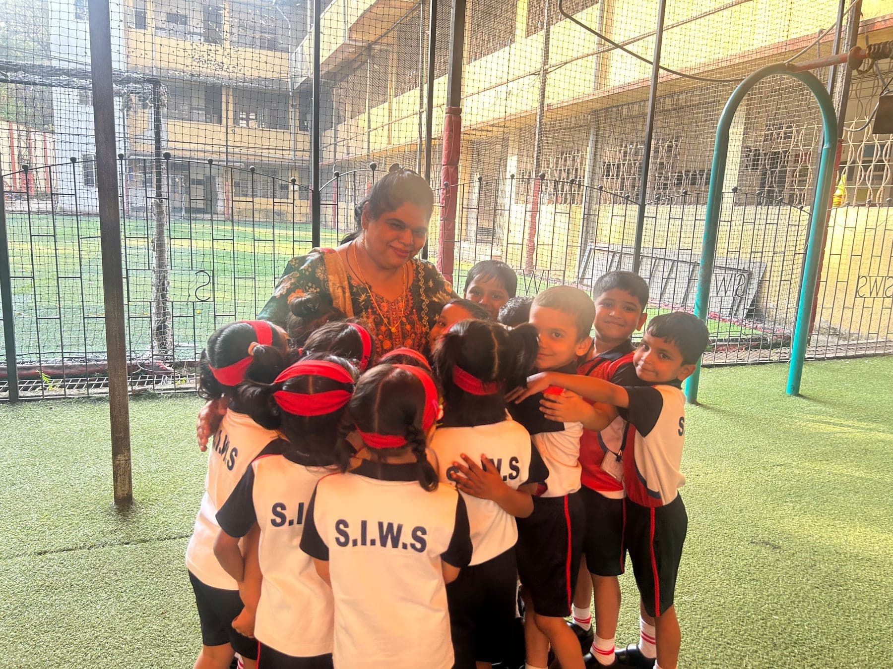 A SIWS teacher surrounded by a group of kindergarten children hugging her on the school play area.