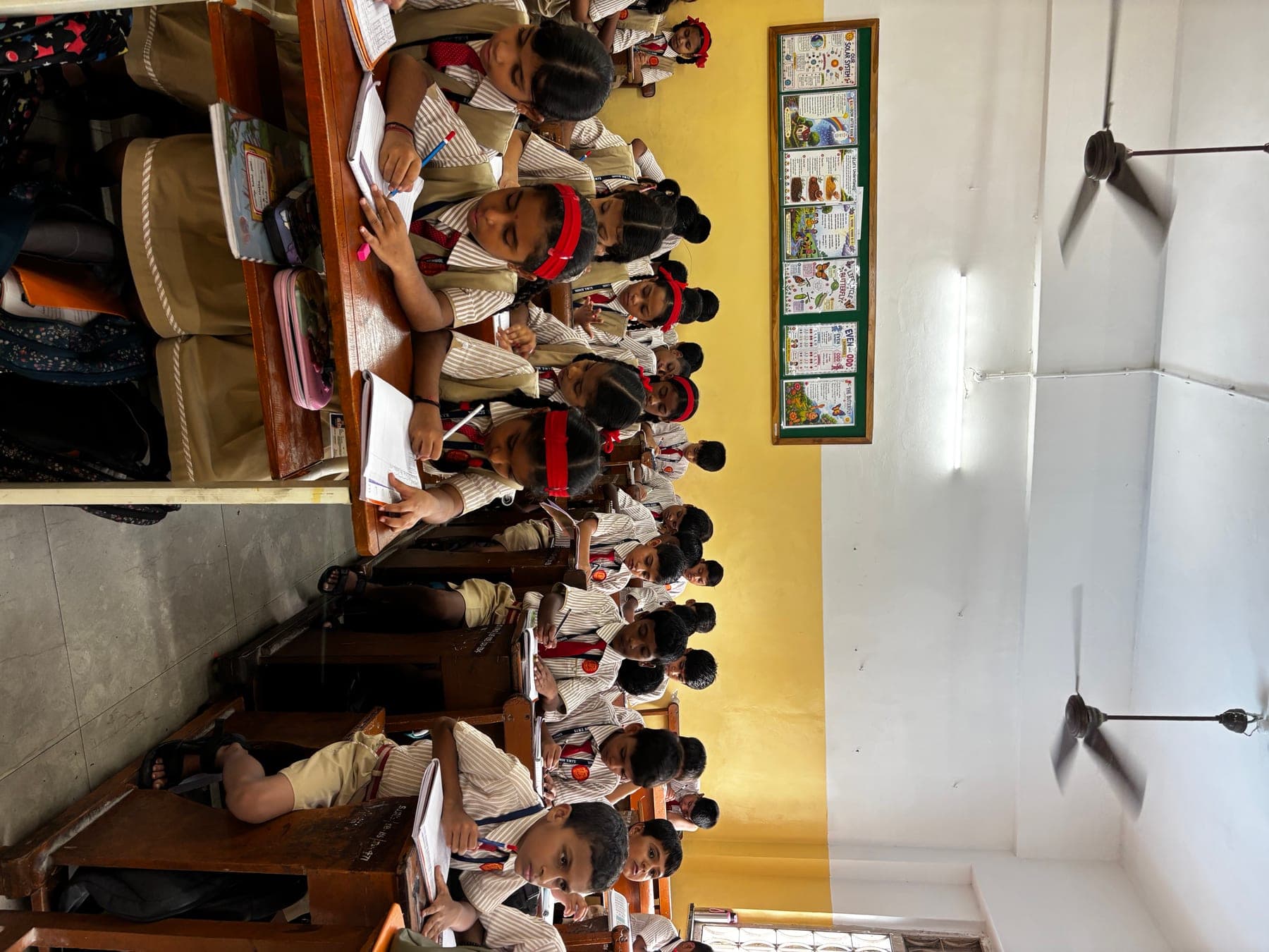 A full Primary class at their bench desks, writing in exercise books, with subject charts on the wall behind them.