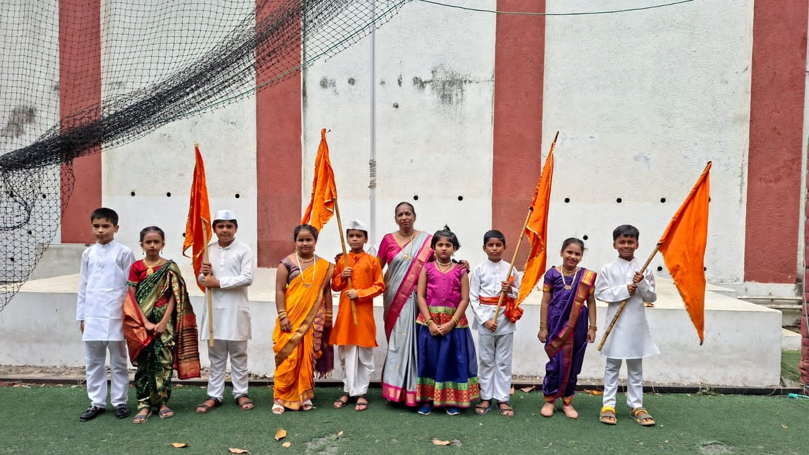 Primary children in traditional Maharashtrian dress holding saffron flags on the school ground, with their teacher beside them.
