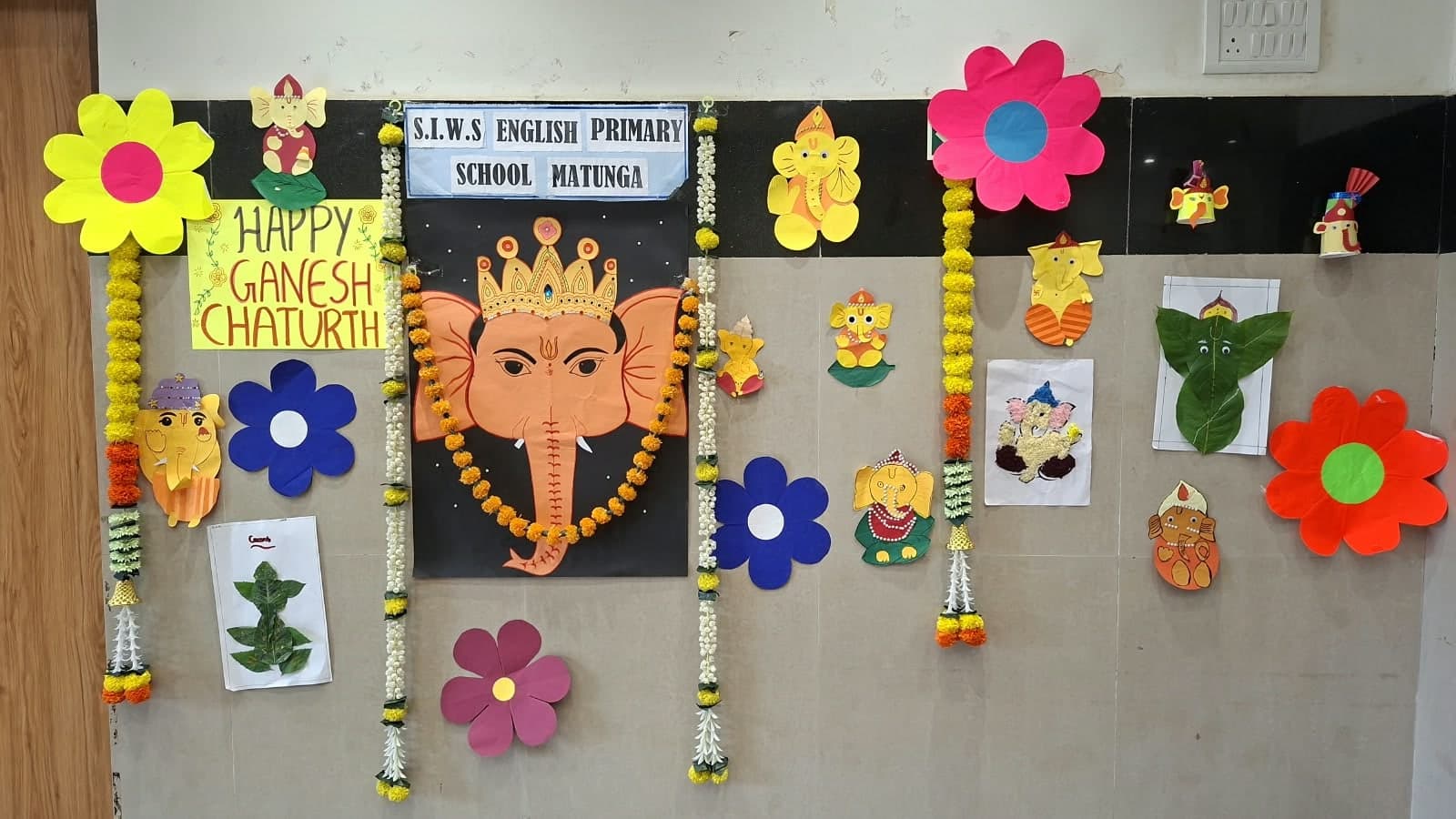 The Ganesh Chaturthi display at S.I.W.S English Primary School, Matunga: a large paper Ganesha face garlanded with marigolds, a “Happy Ganesh Chaturthi” sign, paper flowers and Ganesha crafts on the wall.