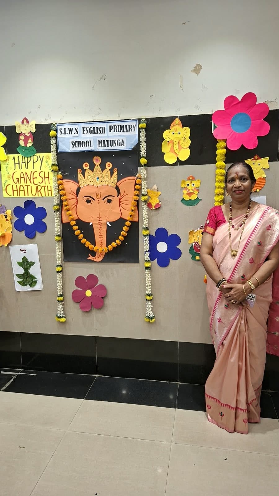 A woman in a peach silk saree standing beside the Ganesh Chaturthi display in the school corridor.