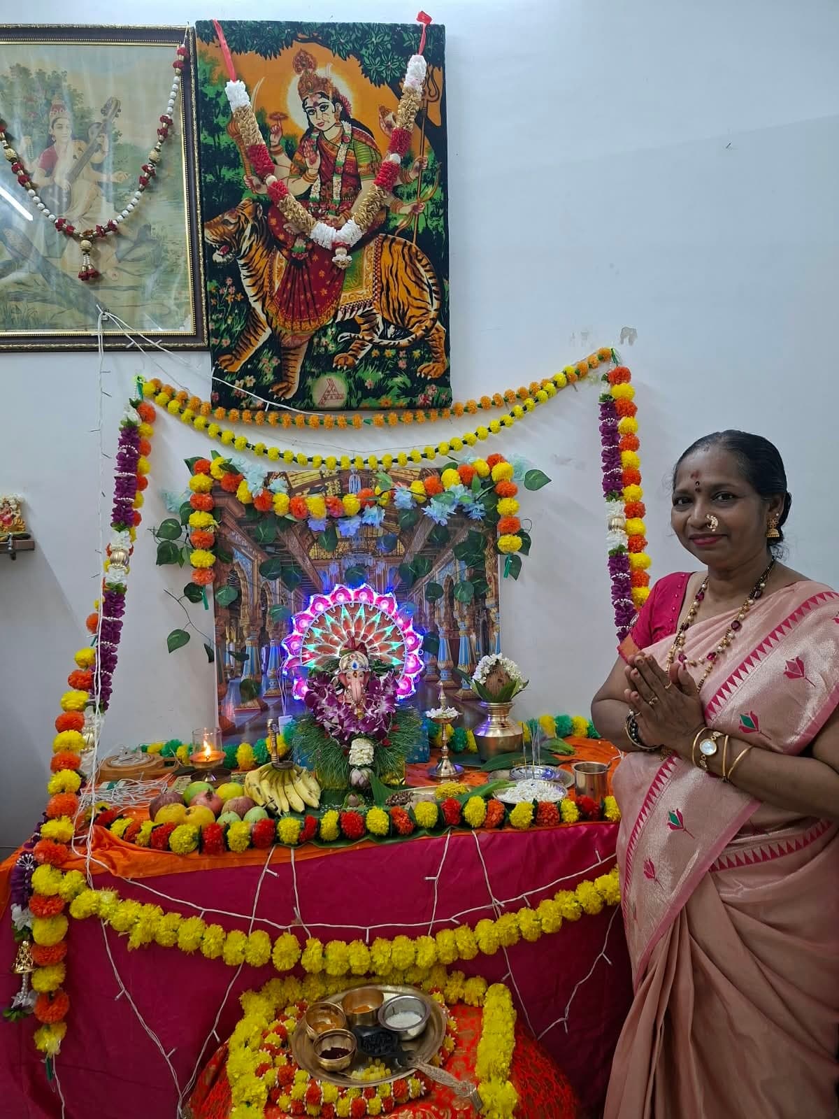A woman in a peach silk saree standing with folded hands beside the decorated Mangala Gauri altar.