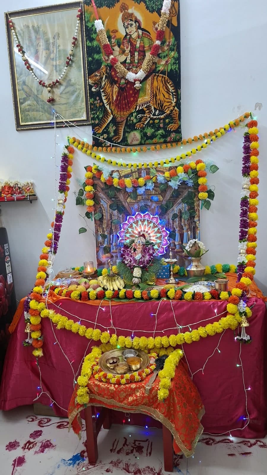 The Mangala Gauri altar: a Ganesha idol garlanded with orchids before a lit backdrop, with fruit, a brass lamp and a kalash on a red-draped table hung with marigold garlands, beneath a painting of Durga on a tiger.