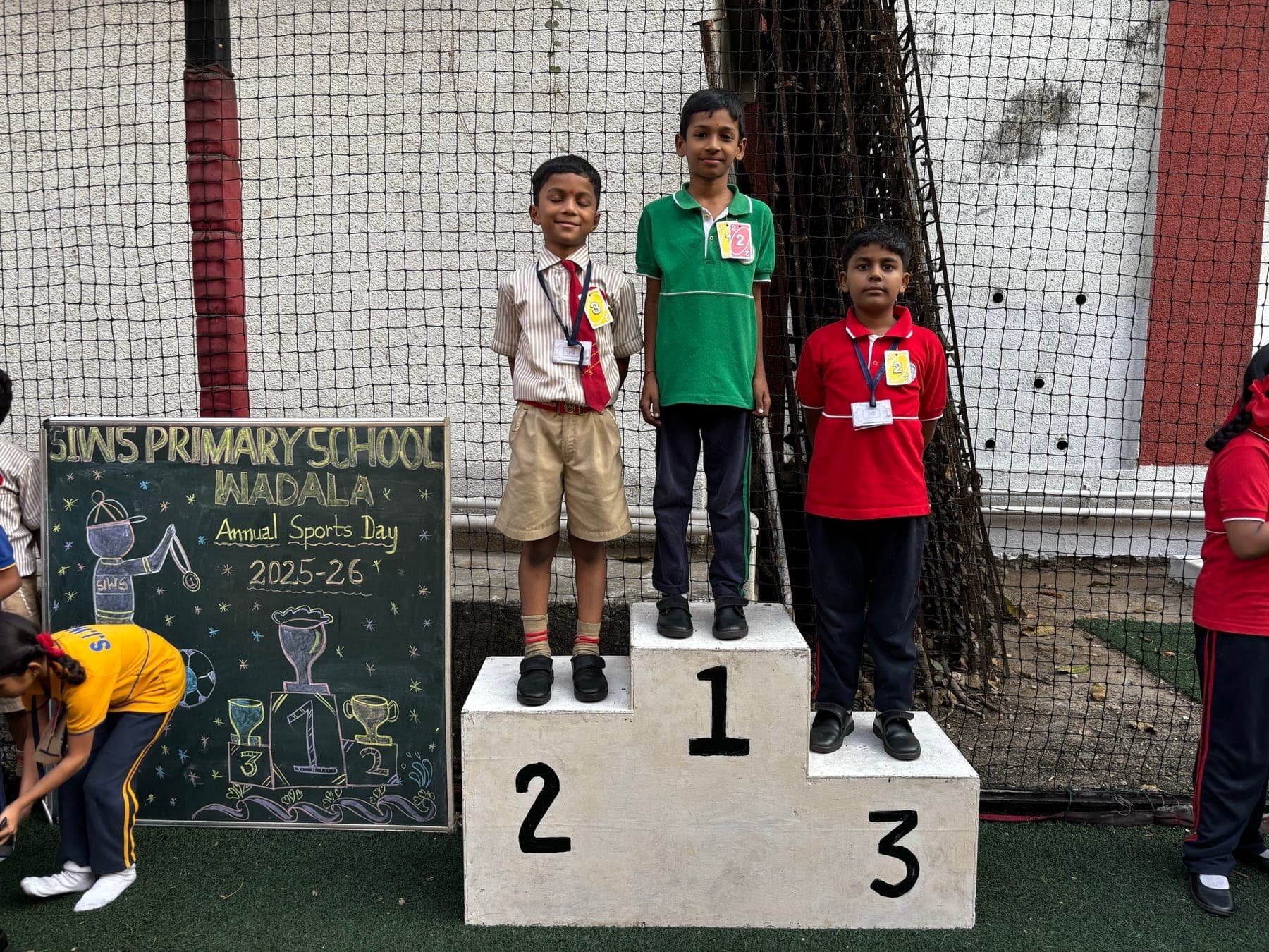 Three Primary boys on the winners’ podium — second in a cream shirt, first in green, third in red — beside the Annual Sports Day 2025-26 chalkboard.