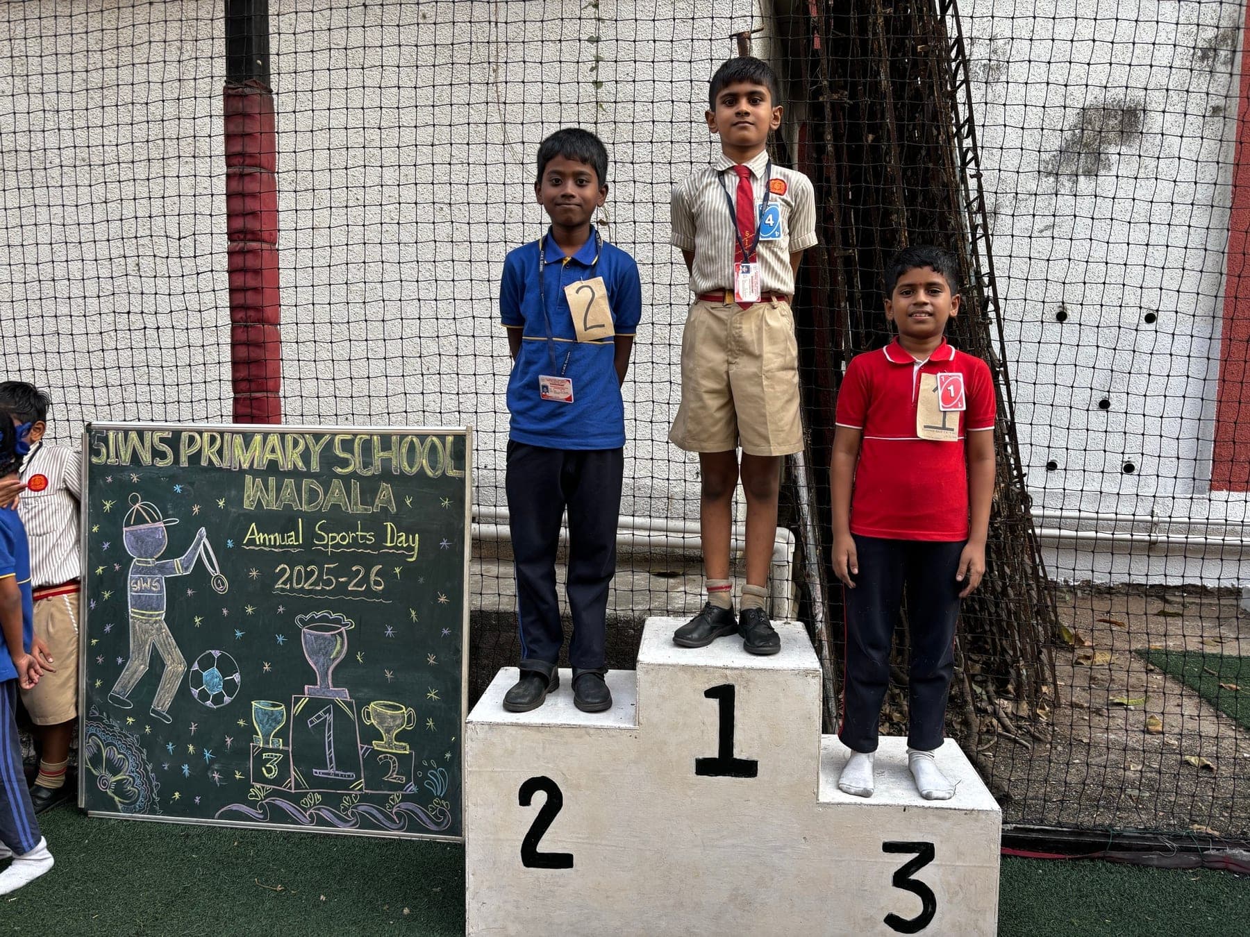 Three Primary boys on the winners’ podium — second in blue, first in a cream shirt, third in red — with the sports day chalkboard and the netting of the turf pitch behind them.