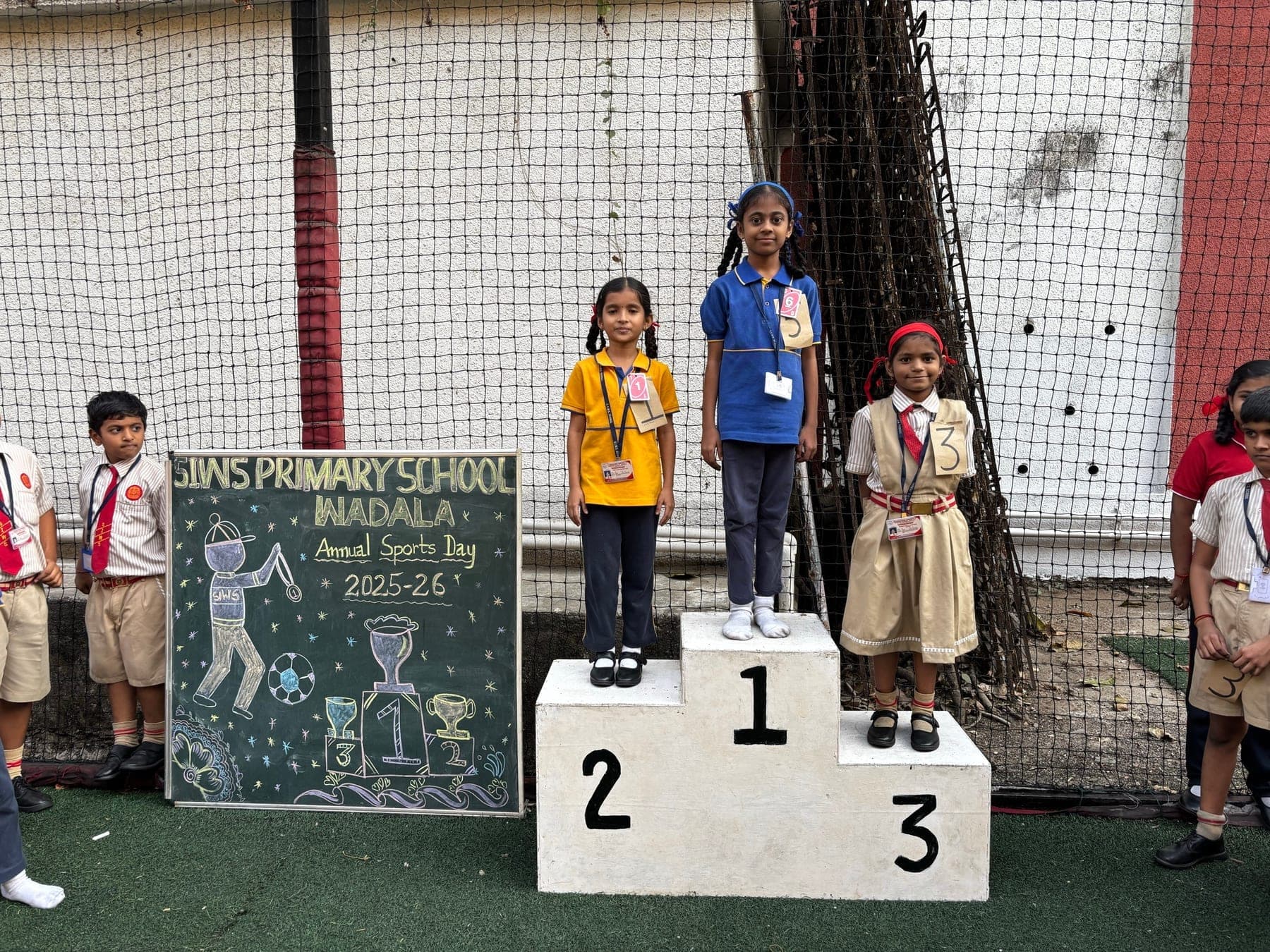 Three Primary children on the winners’ podium — second in yellow, first in blue, third in a cream pinafore with a red headband — beside the Annual Sports Day 2025-26 chalkboard.