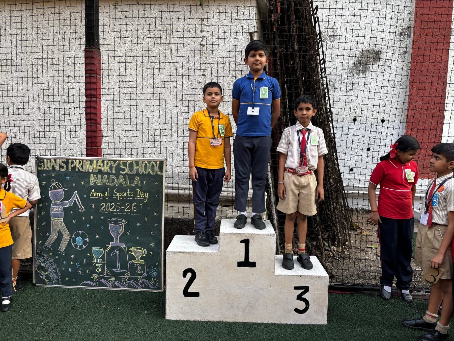 Three Primary boys on the winners’ podium — second in yellow, first in blue, third in a cream shirt and tie — beside the Annual Sports Day 2025-26 chalkboard.