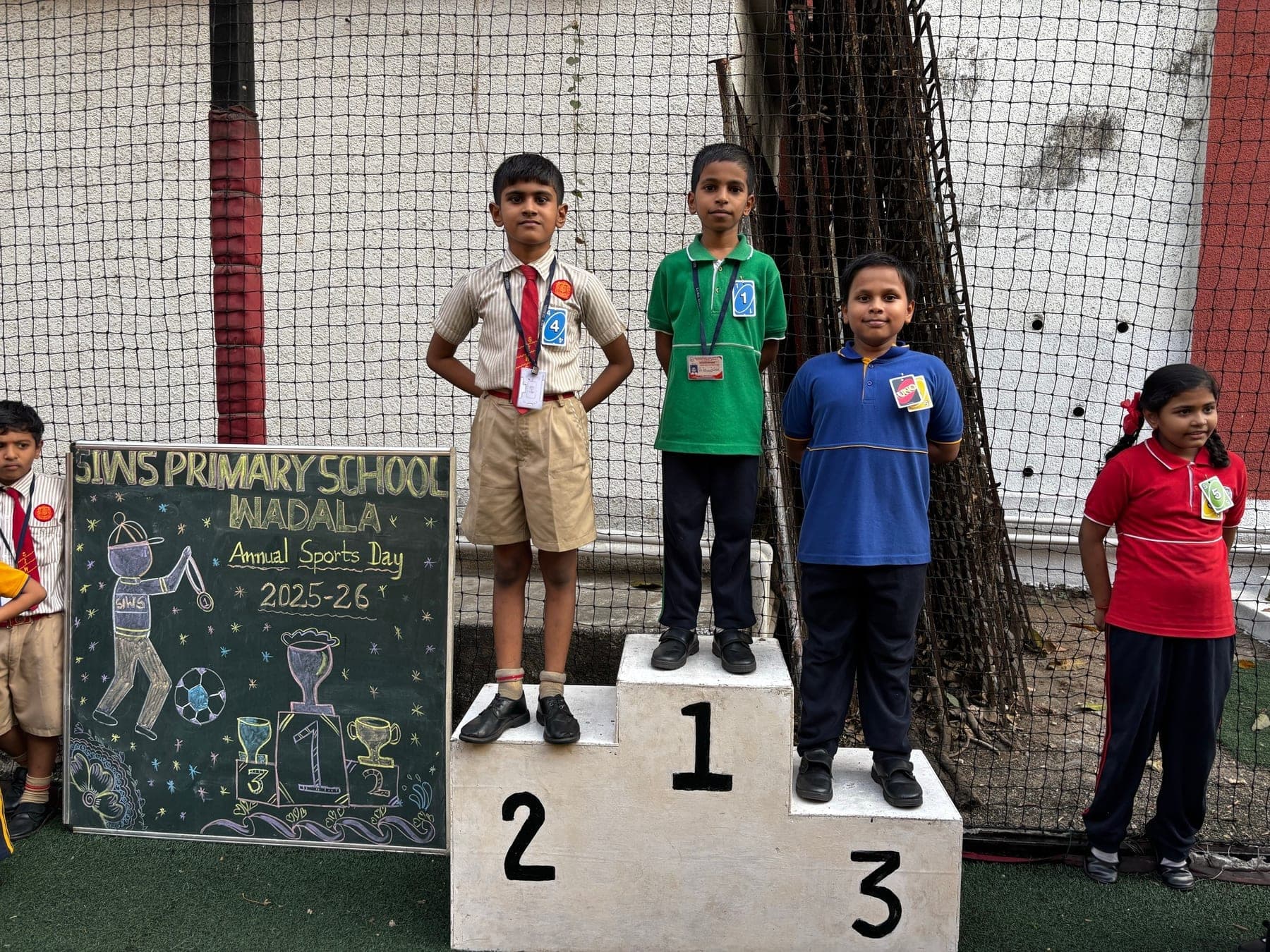 Three Primary boys on the winners’ podium — second in a cream shirt, first in green, third in blue — beside the Annual Sports Day 2025-26 chalkboard.