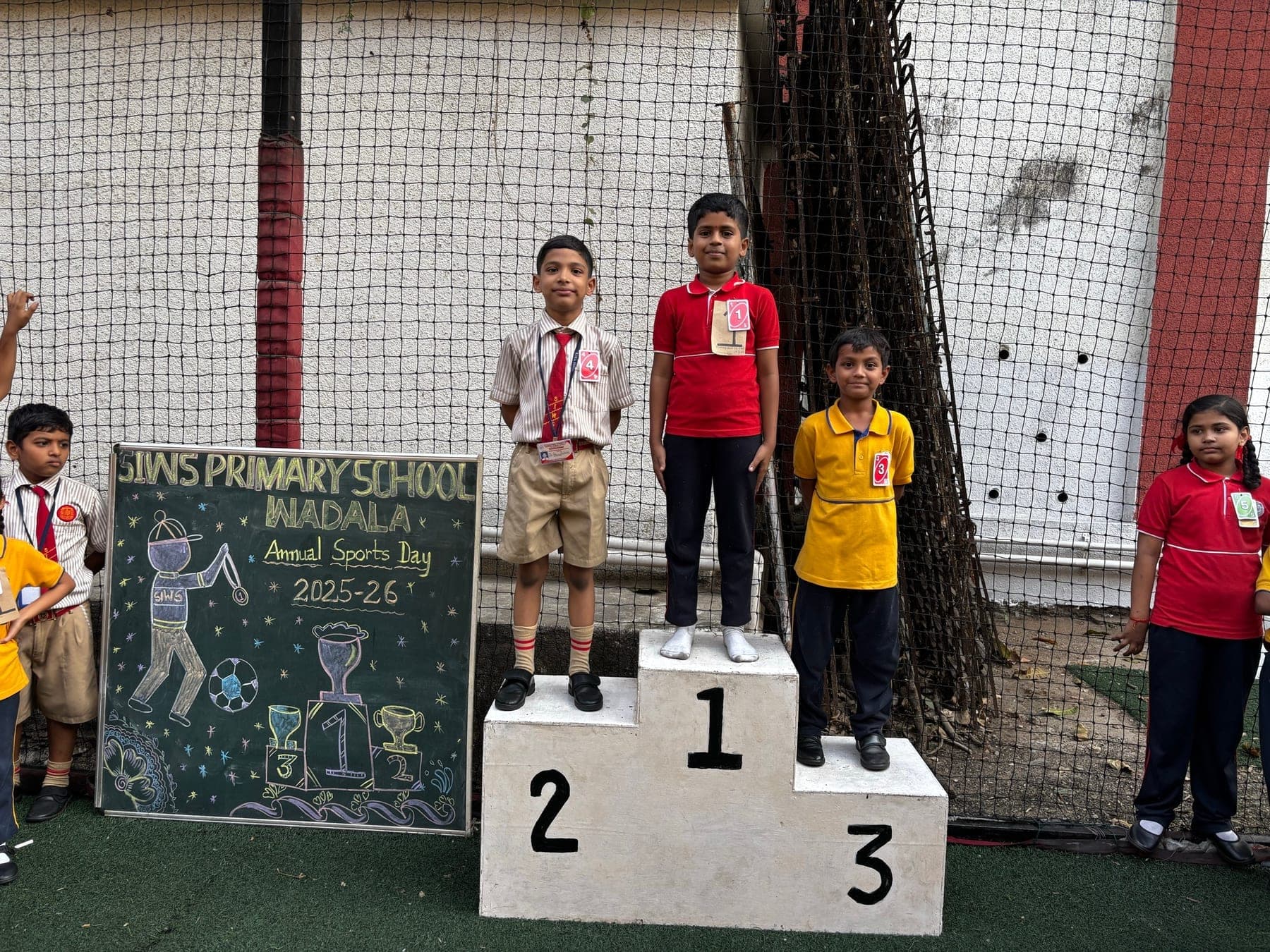Three Primary children on the winners’ podium — second in a cream shirt, first in red, third in yellow — beside the Annual Sports Day 2025-26 chalkboard.