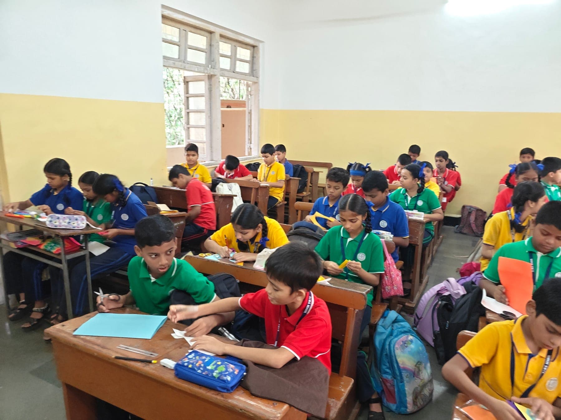 A full Secondary classroom of students in house-colour shirts working with coloured paper and scissors at wooden desks.