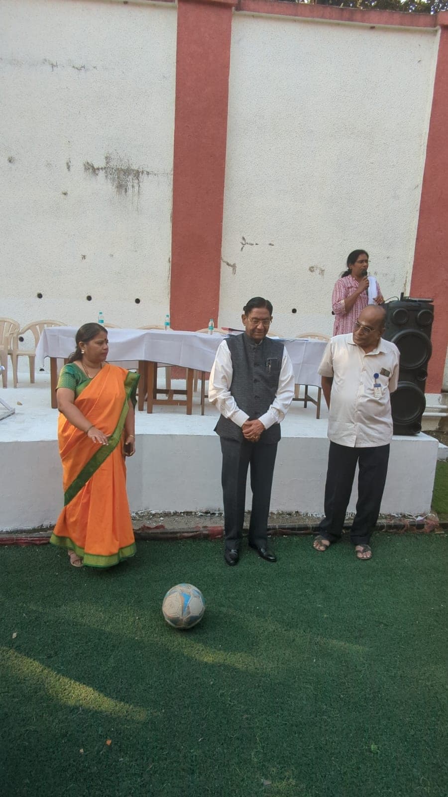 Two men and a woman in a sari standing over a football on the turf pitch beside a draped table, about to start the inter-school football league.