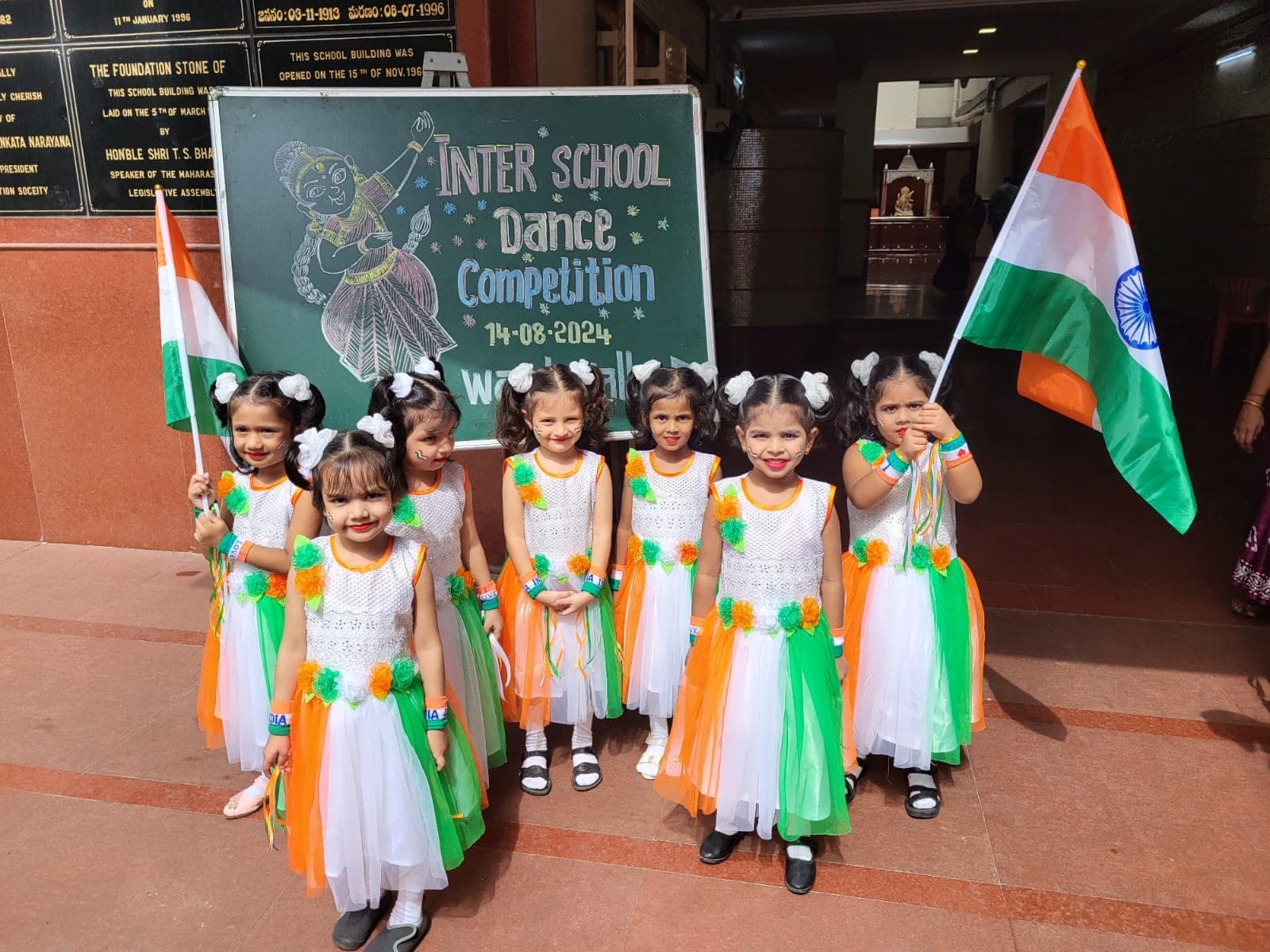 Seven Kindergarten girls in orange, white and green dance costumes holding Indian flags, in front of a blackboard reading “Inter School Dance Competition, 14-08-2024”.