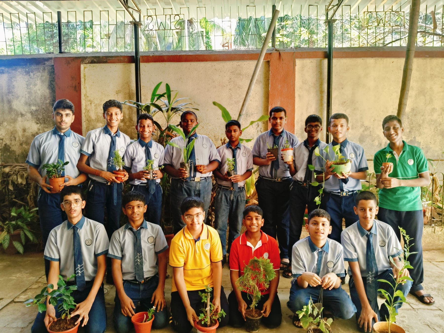 Sixteen Secondary School students in two rows on a school veranda, each holding a potted plant or sapling they have grown.