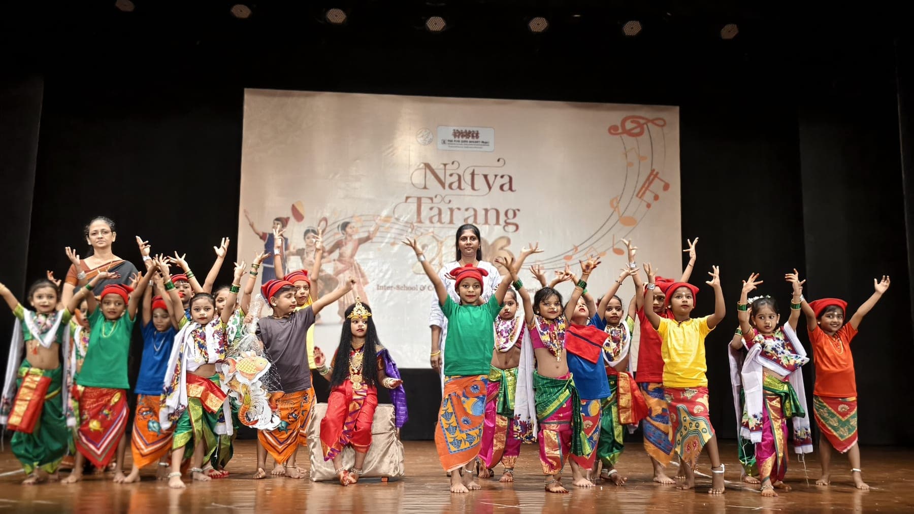A stage full of young SIWS students in bright regional costume, arms raised mid-performance, at the Natya Tarang inter-school dance competition.