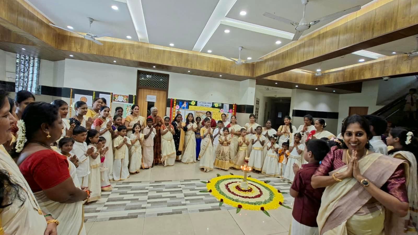 Children and teachers in cream-and-gold Kerala dress standing in a wide circle around a floral pookalam and lit brass lamp in the school hall, hands folded in greeting.