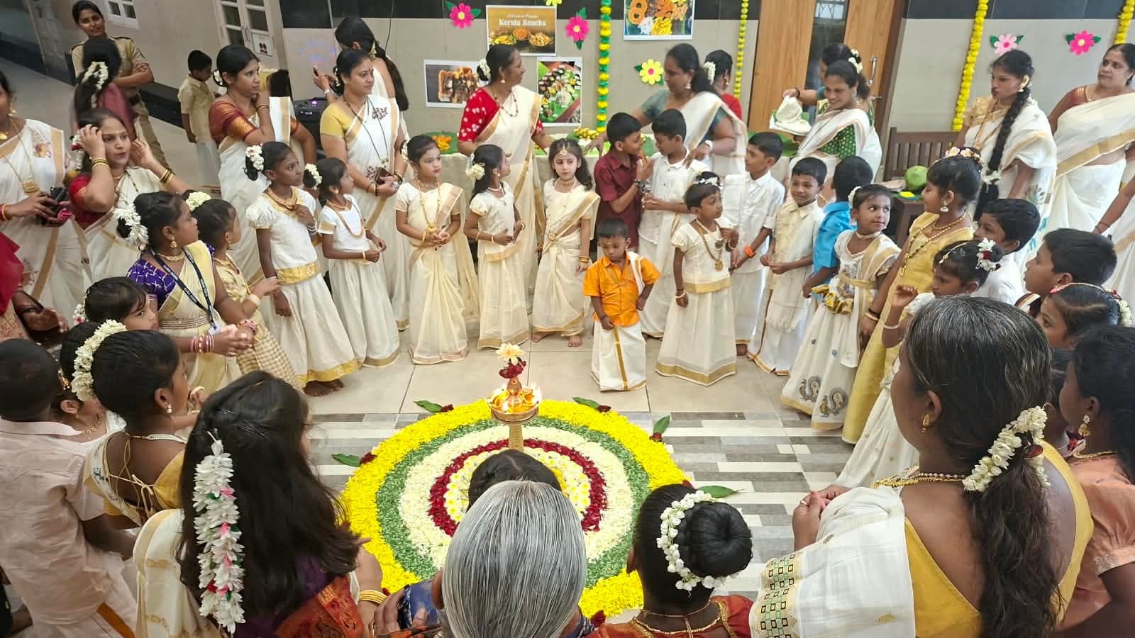 Young children in traditional Kerala dress crowded around a large circular flower pookalam of yellow, white, red and green petals, with a Kerala Snacks display board behind them.