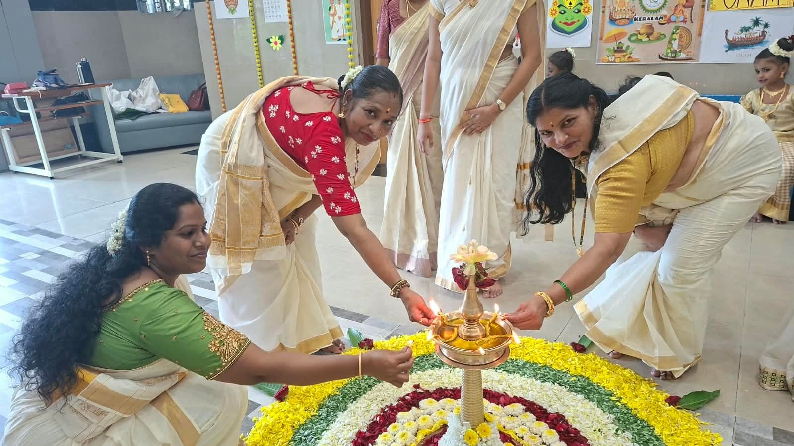 Three teachers in Kerala sarees bending together to light the wicks of a tall brass lamp standing at the centre of the flower pookalam.