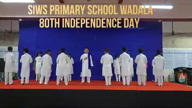 Children in white kurtas standing in a line on a stage under a blue backdrop reading “SIWS Primary School Wadala, 80th Independence Day”.