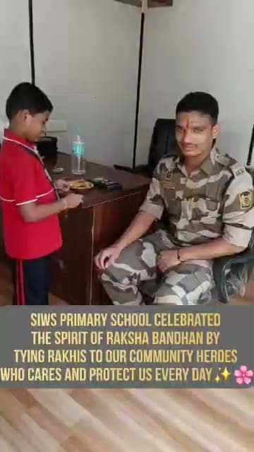 A boy in a red T-shirt tying a rakhi to the wrist of a uniformed serviceman seated at a desk.